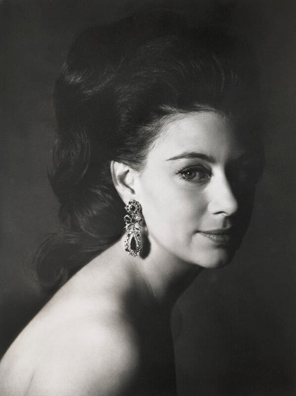 Black and white portrait of a woman with dark, voluminous hair, looking at the camera from a side angle. She wears large, ornate earrings. The background is dark, highlighting her profile and elegant features.
