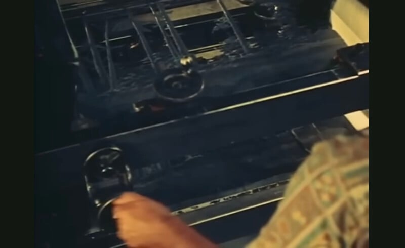A person operates a vintage printing press, turning a handle. The press features metal gears and ink rollers. The individual is wearing a patterned shirt, focusing on the mechanism. The image conveys a sense of traditional printing techniques.