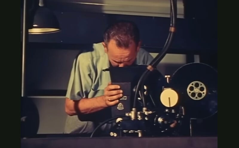 A man looks into a vintage film projector with reels and lenses, focused on his work. The room is dimly lit by an overhead lamp.