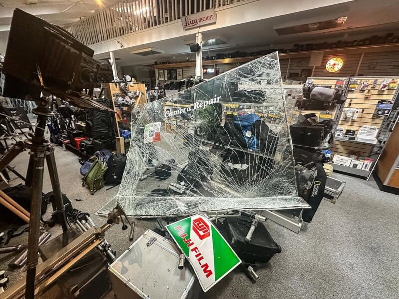 A cluttered camera store with a shattered glass display titled "Camera Repair" prominently featured. Various camera equipment surrounds the display. Signs for Fujifilm are visible amidst the chaos.