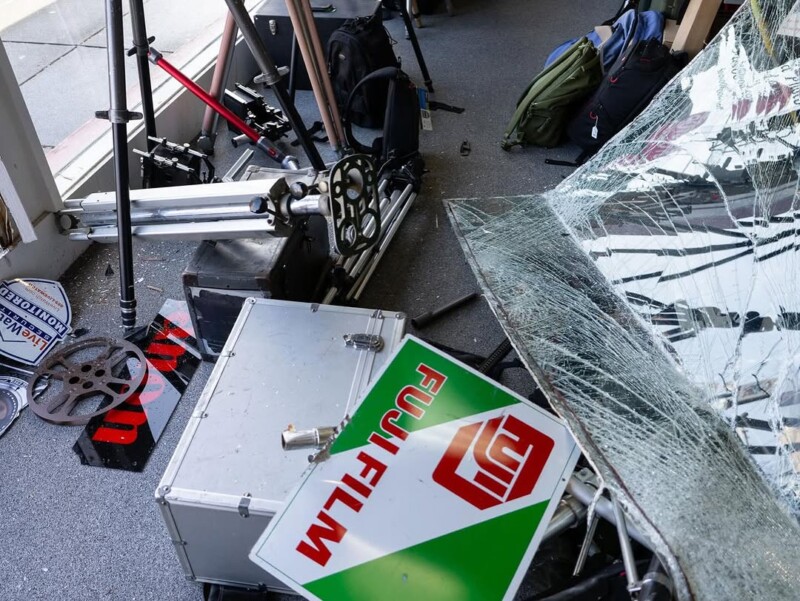 A shattered glass pane lies on a studio floor amid disassembled film equipment, a cracked "Fujifilm" sign, tripods, and a camera dolly. Backpacks are in the corner, and a window can be seen in the background.