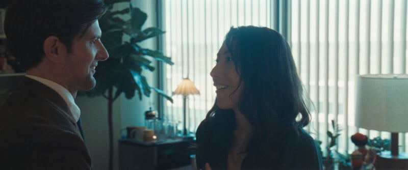 A man and a woman smile at each other in an office with large windows and vertical blinds. There are plants, a lamp, and office items visible in the background.