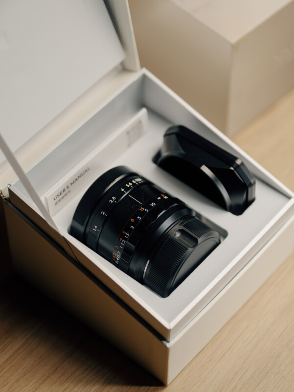 A camera lens and lens hood are neatly placed inside a white presentation box, alongside a user manual, on a wooden surface.