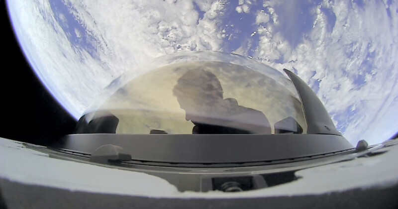 A person is visible through the dome-shaped glass of a spacecraft, with Earth and its cloud formations prominently displayed in the background. The perspective is from space, emphasizing the vastness of the planet below.