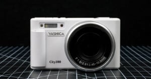 A white Yashica City100 digital camera is displayed upright on a grid-patterned surface against a black background, showing its lens and front details clearly.