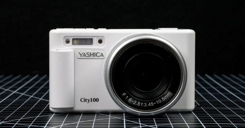 A white Yashica City100 digital camera is displayed upright on a grid-patterned surface against a black background, showing its lens and front details clearly.
