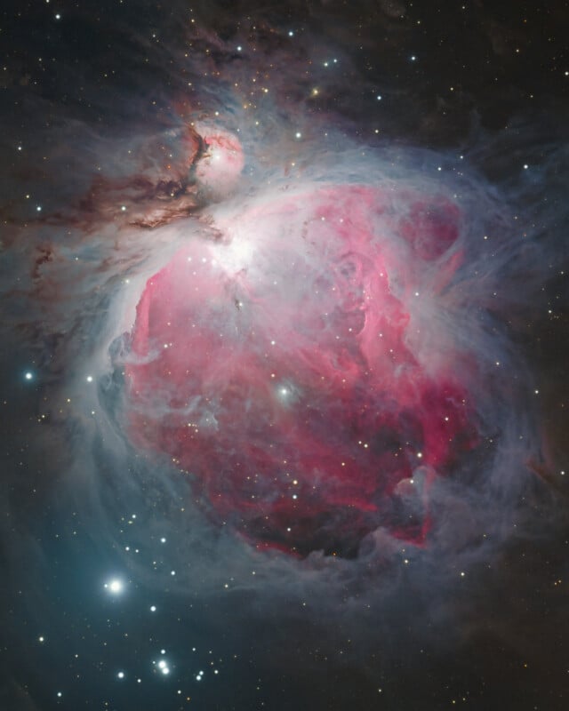 A vibrant image of the Orion Nebula, showing swirling clouds of pink and red gas, bright stars scattered throughout, and a soft glow against the dark space background.