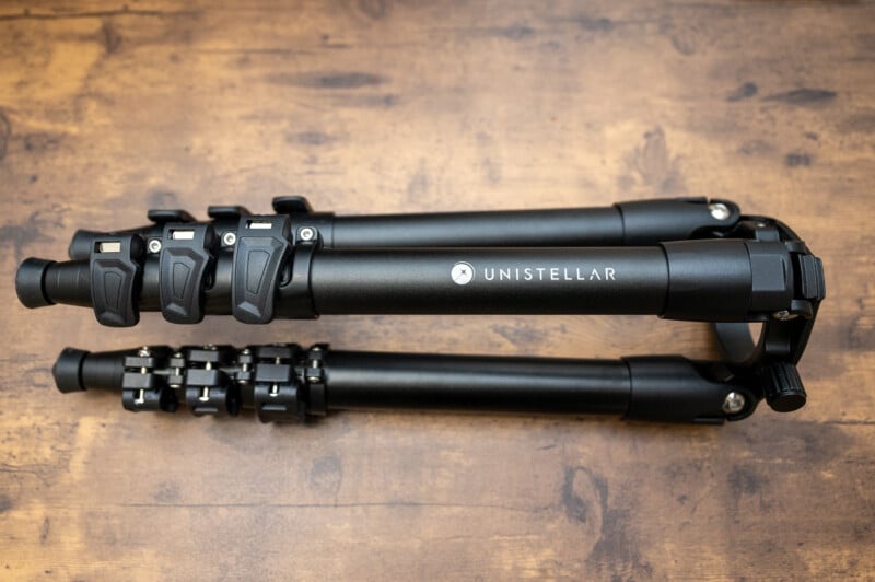 A folded black Unistellar tripod with adjustable leg locks, placed on a wooden surface.