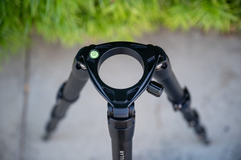 Top-down view of a black tripod with telescoping legs, shown without a center column or camera attached. A bubble level is visible on the tripod plate. Green foliage and gray concrete are in the background.