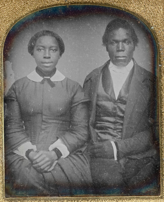 A vintage portrait of a seated couple from the 19th century. The woman wears a dark dress with a white collar, and the man is in a dark suit with a vest and cravat. The image has a textured, aged appearance.