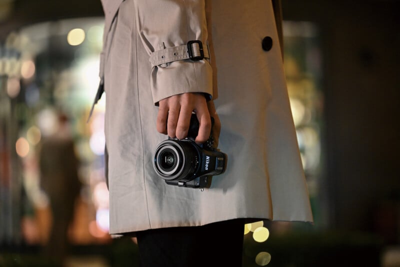A person in a beige trench coat holds a Nikon camera in their hand. The background is blurred with lights and colors, suggesting an urban setting at night.