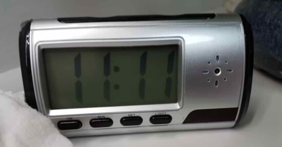A digital clock displays the time 11:11. The clock has a silver frame with buttons labeled "HR," "MIN," "SET," "LIGHT" below the screen. It is placed on a white surface with a partially visible dark object nearby.