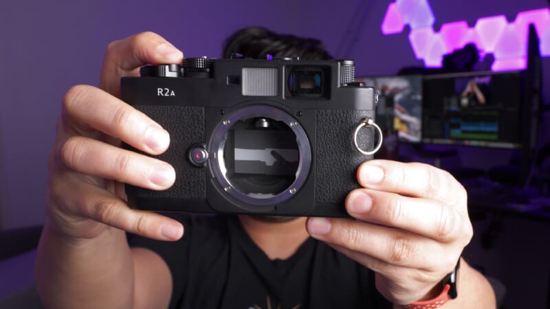 A person holds a large black R2A camera without a lens towards the camera, with both hands visible. The background shows a blurred monitor and glowing geometric lights on the wall.