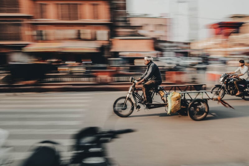 A man rides a modified motorcycle with a trailer attached, carrying bags, through a busy urban street. Motion blur emphasizes speed and activity, with buildings and people in the background.