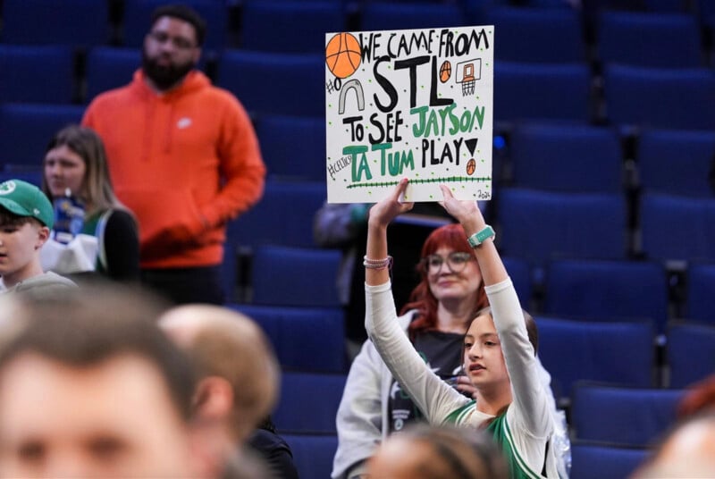 A young fan at a basketball game holds up a sign reading "We came from STL to see Jayson Tatum play" with colorful decorations, standing among other spectators.