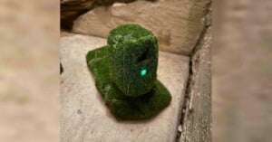 A small sculpture resembling an Easter Island Moai, covered in artificial grass, stands on a concrete surface against a stone wall. A small, glowing green light is visible on its face.