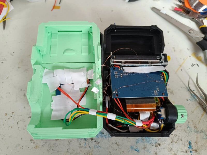 An open electronic device with a green and black casing. Inside are multicolored wires, circuits, and a blue circuit board. The device is placed on a white surface with tools around it.