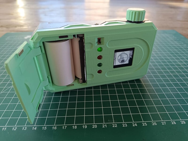 A green 3D-printed camera is placed on a cutting mat. The back is open, showing a roll of white paper inside. Three indicator lights and a small display screen are visible on the back of the camera.