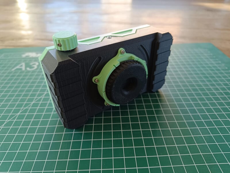 A 3D-printed camera featuring a mint green and black design with a prominent lens and textured grip rests on a green cutting mat with measurement markings. The light wood background adds contrast.