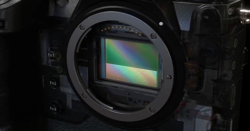 Close-up view of a digital camera body with the lens removed, revealing the camera sensor displaying a rainbow-like reflection. Several function buttons are visible on the left side of the camera.