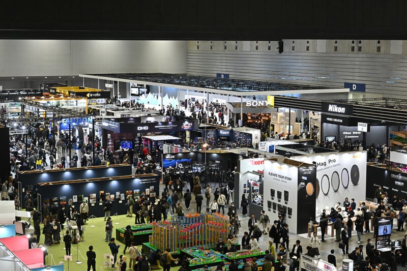 A large, busy trade show floor with many people walking among booths and displays from various camera and tech brands, including Sony and Nikon, inside a spacious convention center.