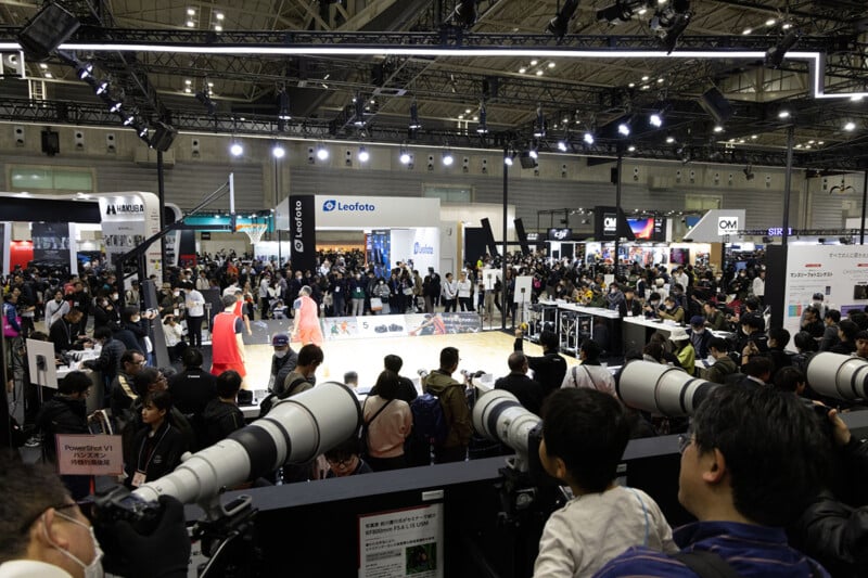 A large crowd attends a photography convention with people observing and photographing models on a central stage; many large telephoto camera lenses are visible in the foreground.