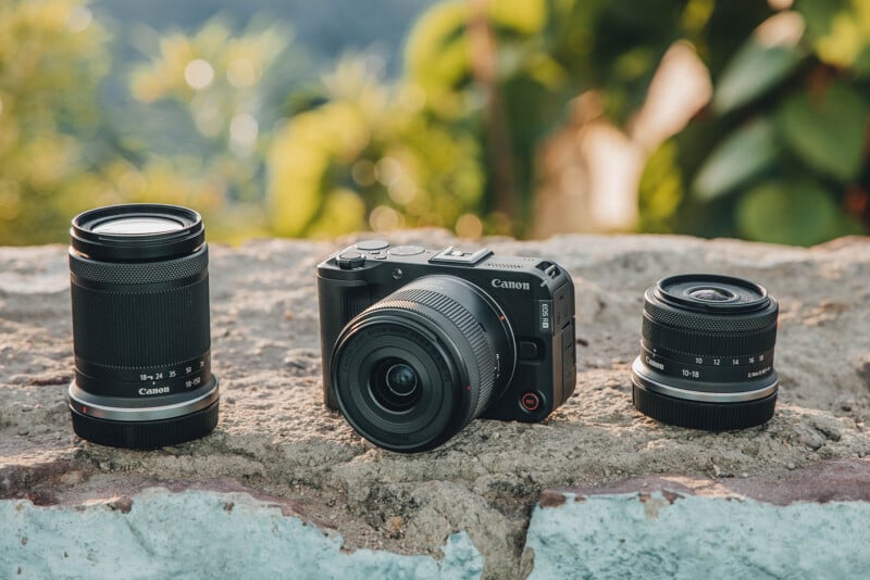 A Canon mirrorless camera with a lens attached is placed on a stone surface outdoors, flanked by two additional interchangeable Canon lenses, with greenery in the blurred background.