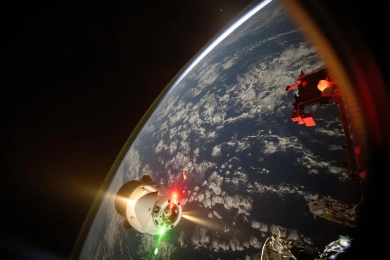 A spacecraft approaches Earth’s atmosphere, illuminated by green and red lights, with clouds and the curve of the planet visible below, and part of a space station structure in the foreground.