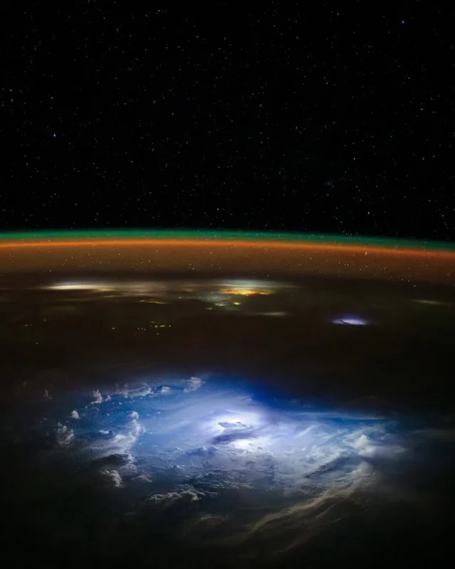 View of Earth’s atmosphere from space, showing glowing clouds with flashes of lightning, a thin orange and green atmospheric layer, and a star-filled black sky above.