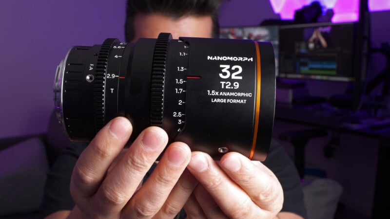 A person holds a Nanomorph 32mm T2.9 1.5x anamorphic large format lens with both hands, in a room with a blurred background featuring a desk, computer monitors, and purple lighting.