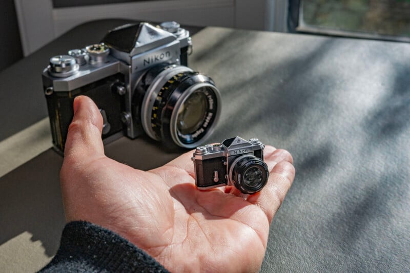A hand holds a miniature model of a Nikon camera, with a full-sized Nikon camera in the background on a table. Sunlight filters through a nearby window, casting soft shadows.