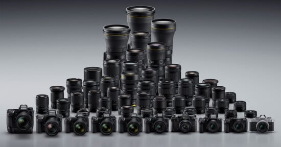 A large collection of digital cameras and interchangeable lenses are arranged in rows, forming a triangular shape, with larger lenses at the back and smaller cameras and lenses at the front on a gray background.