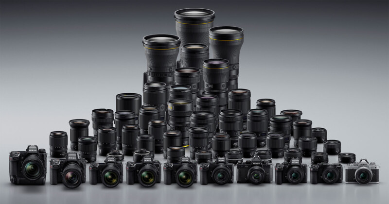 A large collection of digital cameras and interchangeable lenses are arranged in rows, forming a triangular shape, with larger lenses at the back and smaller cameras and lenses at the front on a gray background.