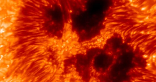 A close-up image of the Sun’s surface showing dark sunspots surrounded by swirling, bright orange and yellow plasma patterns. The contrast highlights the intense heat and energy of the solar activity.