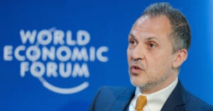 A man in a suit and yellow tie speaks in front of a blue background with the World Economic Forum logo.