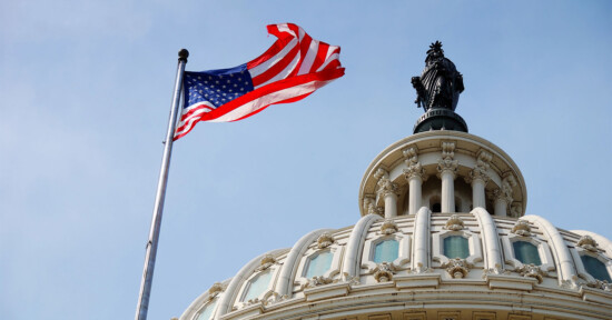 An American flag waves in the wind next to the dome of the U.S. Capitol building, topped by the Statue of Freedom, against a blue sky.