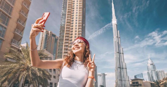 A smiling woman wearing sunglasses and a headband takes a selfie and makes a peace sign, with tall modern buildings and the Burj Khalifa visible in the background under a clear blue sky.