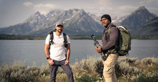 Two men wearing backpacks stand by a lake with tall grass in the foreground and mountains in the background. One holds a camera tripod. Both are dressed for hiking and look toward the camera under a cloudy sky.