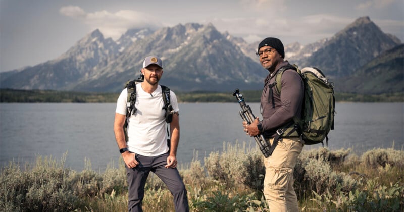 Two men wearing backpacks stand by a lake with tall grass in the foreground and mountains in the background. One holds a camera tripod. Both are dressed for hiking and look toward the camera under a cloudy sky.
