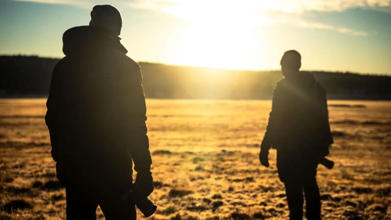 Two people in dark jackets stand in a sunlit open field at sunrise or sunset, with long shadows cast on the golden grass and a bright sky in the background.