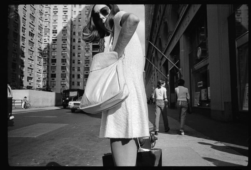 A woman wearing sunglasses and a dress stands on a city sidewalk, rummaging through a large bag. Tall buildings and pedestrians are visible in the background. The image is in black and white.