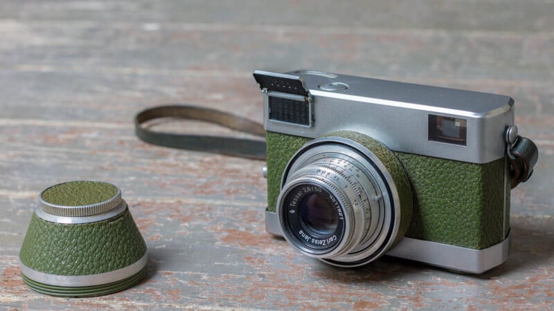 A vintage camera with green leather accents sits on a weathered wooden surface beside a matching green and silver lens cap. The camera has a large lens and a brown wrist strap attached.