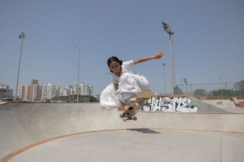 A young girl wearing a white dress performs a skateboard trick in a skate park, mid-air above a ramp, with buildings and graffiti visible in the background under a clear sky.