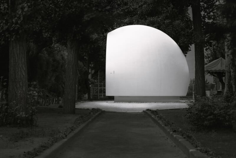 A large, white, minimalist half-sphere structure sits on a circular base in a park, surrounded by trees and shaded paths, with a tent visible in the background.