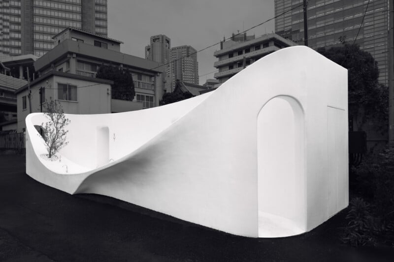 A modern, minimalist public restroom with smooth, curving white walls stands in an urban area surrounded by buildings and skyscrapers. The structure features arched doorways and a small tree planted inside.