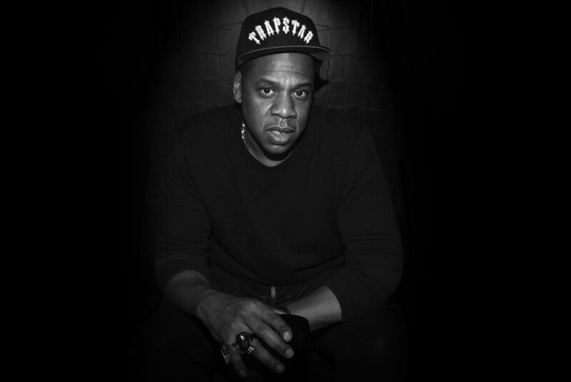 A man wearing a "TRAPSTAR" cap and a dark sweater sits against a black background with a serious expression, holding an object in his hand. The image is in black and white.