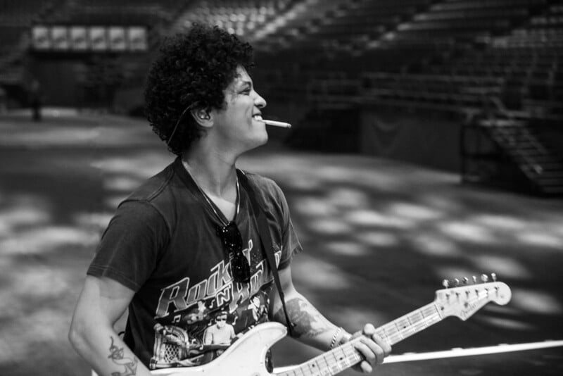 A musician with curly hair smiles while playing an electric guitar in an empty arena, wearing a t-shirt and sunglasses with a cigarette in his mouth.