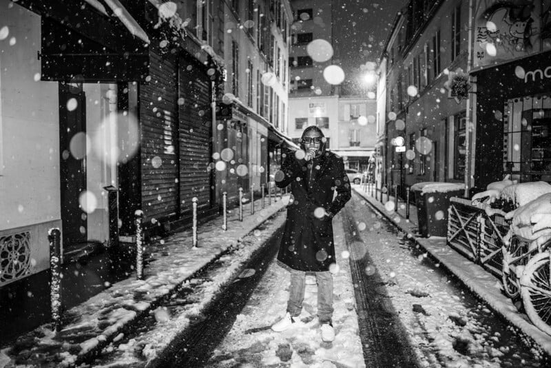 A person in a long coat stands in the middle of a snowy city street at night, snowflakes falling heavily around them. The street is lined with buildings and bicycles, and illuminated by streetlights.