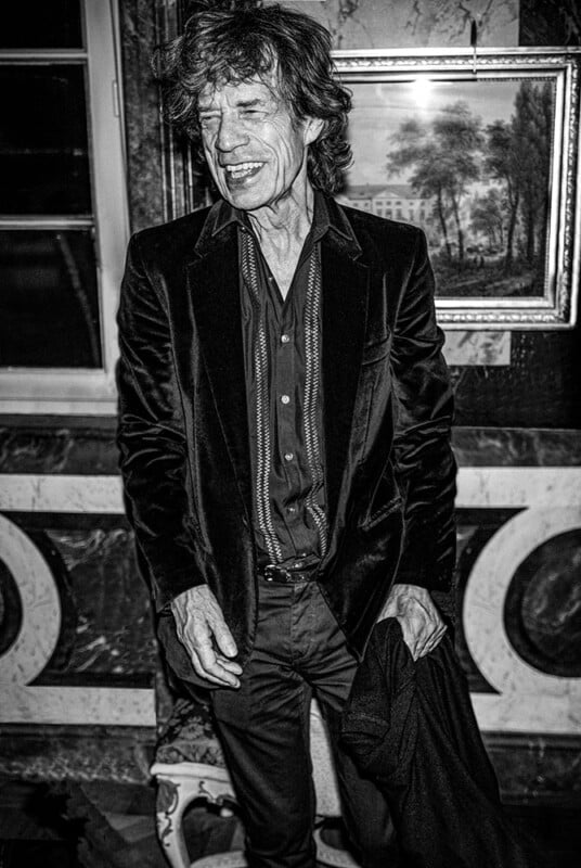 A man in a dark velvet suit stands indoors, smiling with eyes closed. The background features a framed landscape painting and ornate wall decor. The photo is in black and white.