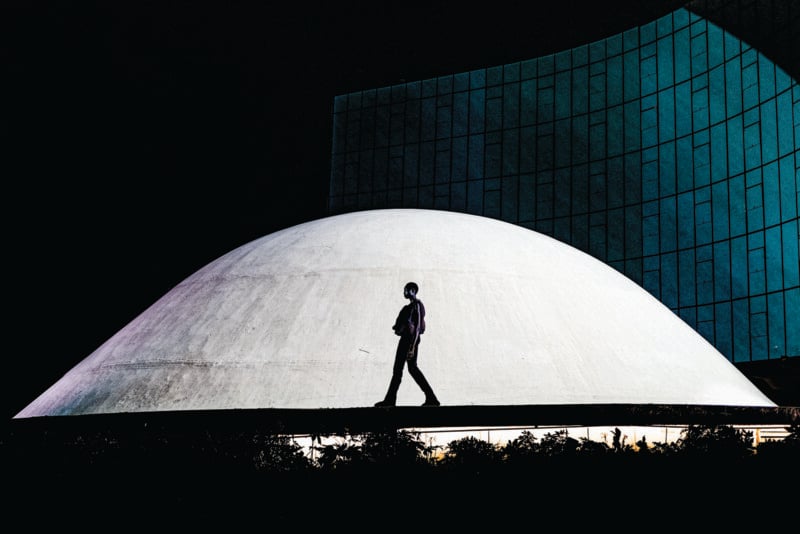 A person walks in front of a large, white dome-shaped structure at night, with a modern glass building in the background and dramatic contrast between light and shadow.